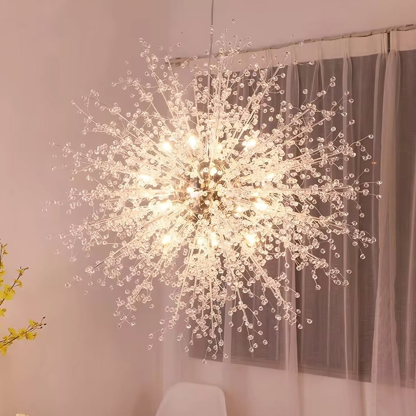 An Astra 35" Spark Ball Pendant Lighting Fixture from Morsale.com, resembling a burst of fireworks, hangs in a room with light pink walls. Below it, a vase with yellow flowers is placed on a white table that also has some other flowers. A white chair is positioned next to the table by a window with sheer curtains, illuminated by 3W G4 LED bulbs.