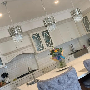 A modern kitchen with white cabinetry, a white island countertop, and three Gio Crystal Kitchen Light Fixtures by Morsale.com hanging above. The backsplash is made of white subway tiles. A vase with colorful flowers sits on the island, and two gray upholstered chairs are placed at the counter.