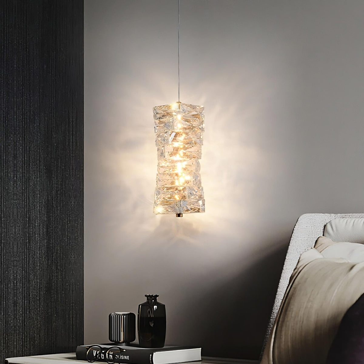 A modern, cylindrical **Bacci Crystal Pendant Light Fixture** from **Morsale.com** hangs from the ceiling, casting a warm glow against a grey wall. Below the light, a side table holds a black vase and several stacked books. Part of a cushioned chair is visible on the right side of the image.