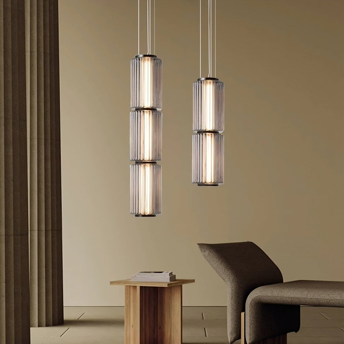 A modern minimalist interior featuring three vertically hanging Steve Kano Glass Pendant Light Fixtures from Morsale.com, each with two cylindrical shades incorporating energy-efficient LED technology. Below, a wooden side table with a book stands beside a sleek, curved chair against a beige backdrop.