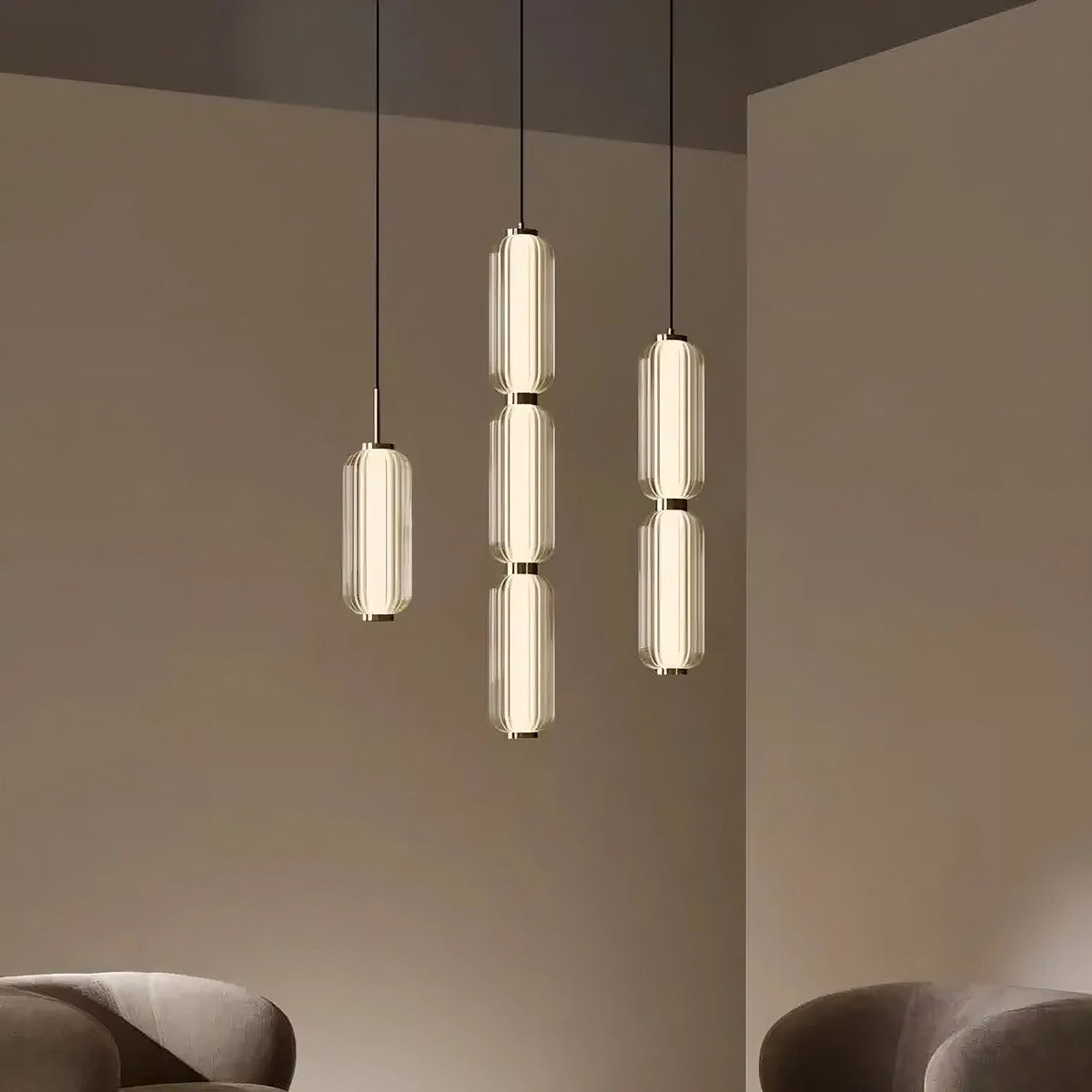 A minimalist room features three vertically aligned Modern Minimalist LED Pendant Light Fixture by Morsale.com with elongated, ribbed glass shades hanging from the ceiling. Two modern, curved beige armchairs are positioned in the foreground, complementing a neutral and calming setting enhanced by subtle touches of natural marble.