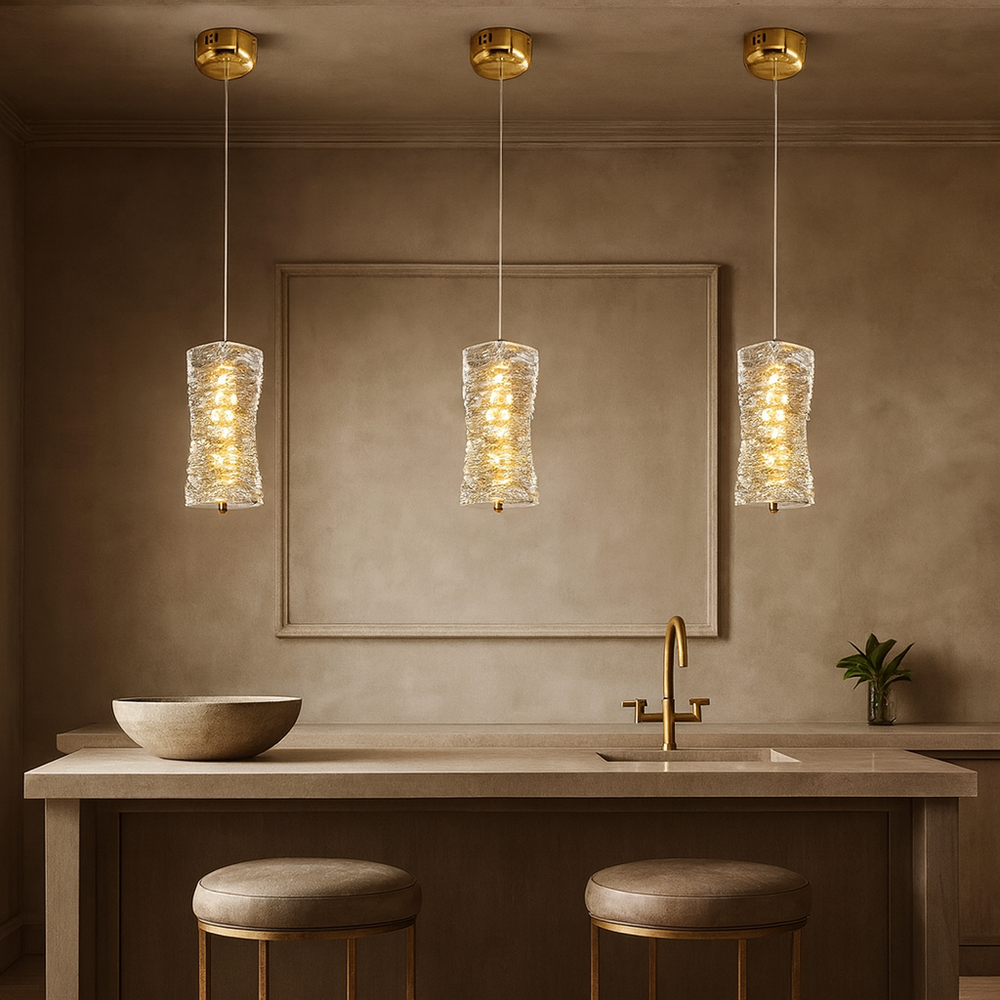 A modern kitchen with a beige island, two round stools, a gold faucet, a bowl on the counter, and three Bacci Crystal Pendants by Morsale.com hanging above. The background has minimalist square wall art and a small plant.