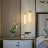 A modern bedroom showcases a sleek nightstand with two white drawers, elegantly adorned with a vase of flowers and a small decorative item. Overhead, the Moonshade Natural Marble Pendant Light Fixtures from Morsale.com hang, casting a warm glow. The neatly made bed with gray and white linens is partially visible.