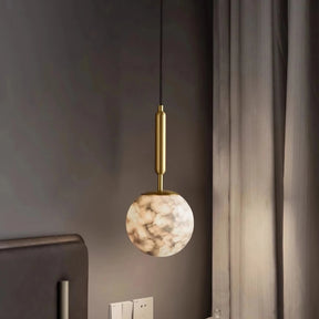 The Moonshade Natural Marble Ball Pendant Light by Morsale, showcasing a modern style with a brass fixture, elegantly hangs beside a bed framed by gray curtains. The dark wall accentuates its charm, while LED bulbs softly illuminate the space. A pair of electrical outlets sit just below this stylish centerpiece.