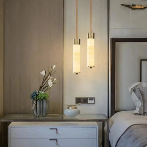 A modern bedroom showcases a sleek nightstand with two white drawers, elegantly adorned with a vase of flowers and a small decorative item. Overhead, the Moonshade Natural Marble Pendant Light Fixtures from Morsale.com hang, casting a warm glow. The neatly made bed with gray and white linens is partially visible.