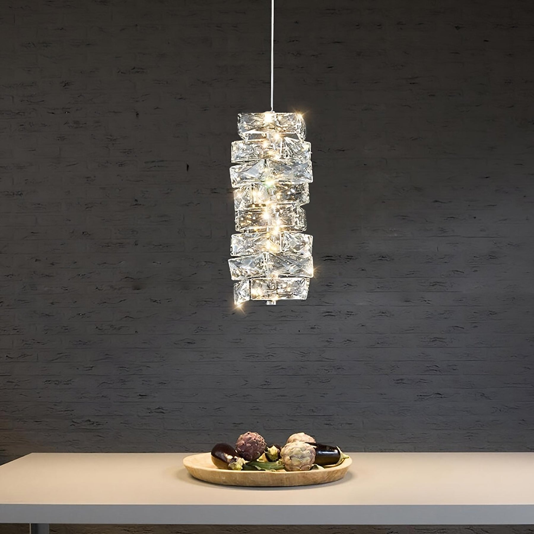 A modern, rectangular Bacci Crystal Pendant Light Fixture by Morsale.com featuring a geometric glass design and handmade crystals hangs from the ceiling over a minimalist table. On the table is a round wooden tray holding various vegetables, including eggplants and artichokes, set against a dark brick wall background.