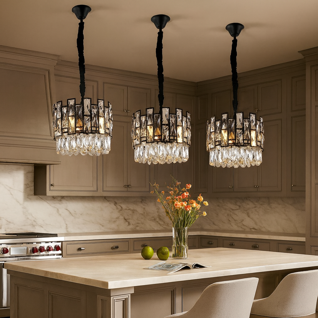 Three Molini Crystal Lights from Morsale.com with shimmering accents hang above a beige kitchen island holding a vase of flowers, two green apples, and an open book. The kitchen features taupe cabinets and a marble backsplash.