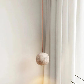 The Travertine Ball Pendant Light Fixture from Morsale.com, crafted from natural travertine stone, features a beige, porous surface that exudes a rustic and organic charm as it hangs from the ceiling. This elegant light fixture is suspended by a brown cord against a plain, off-white background.