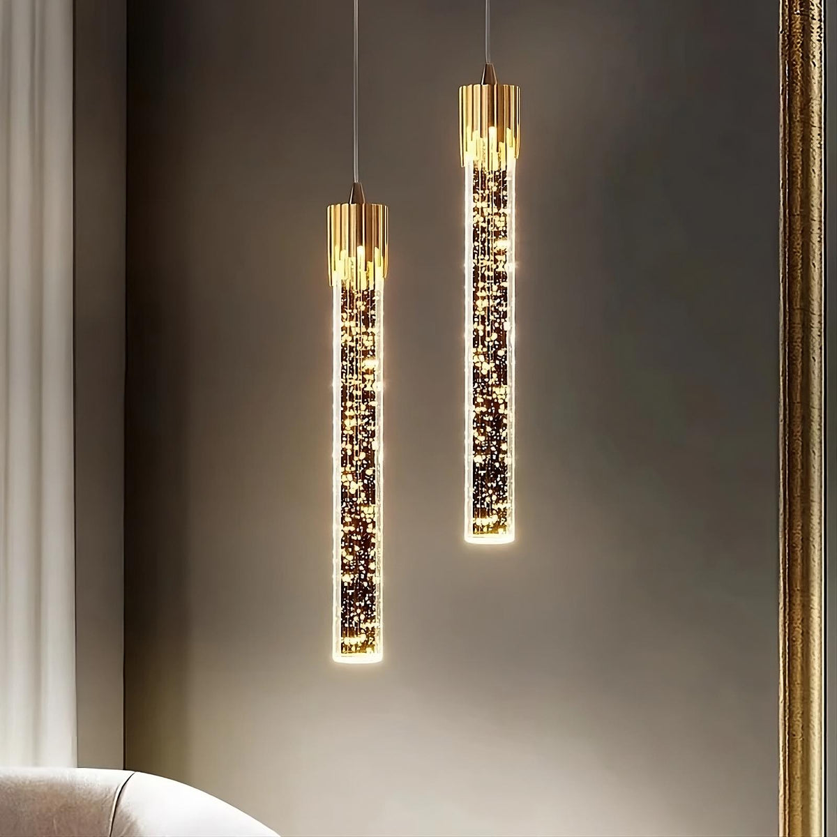 Two modern, cylindrical pendant lights with gold tops and sparkling, crystal-encrusted bodies hang from the ceiling in front of a beige wall. These Modern Minimalist LED Pendant Lights by Morsale.com feature 3W LED Chips and a frosted exterior. A partial view of a white sofa and a gold-edged mirror frame are also visible in the scene.