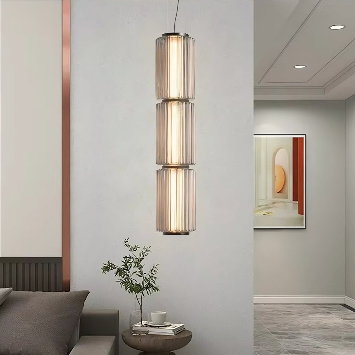 A modern interior room with a sleek Steve Kano Glass Pendant Light Fixture from Morsale.com featuring energy-efficient LED technology hanging from the ceiling. A small round table with a potted plant and cup is placed against the wall under the light. A framed abstract artwork hangs on the wall in the background.