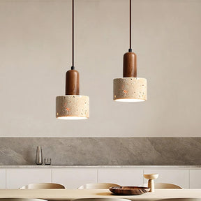 A minimalist kitchen features two Morsale.com Natural Travertine Pendant Lamps with wooden tops and stone-like concrete shades, illuminated by LED bulbs, hanging above a light wood table with a shallow wooden bowl. The background includes a marble backsplash and a counter with minimal décor.
