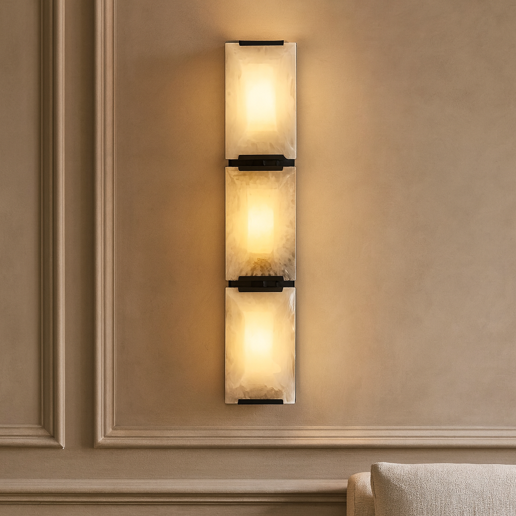 The Brookhill Calcite Crystal Sconce from Morsale.com, featuring three square frosted glass lights, is mounted vertically on a beige paneled wall beside a light-colored upholstered sofa.