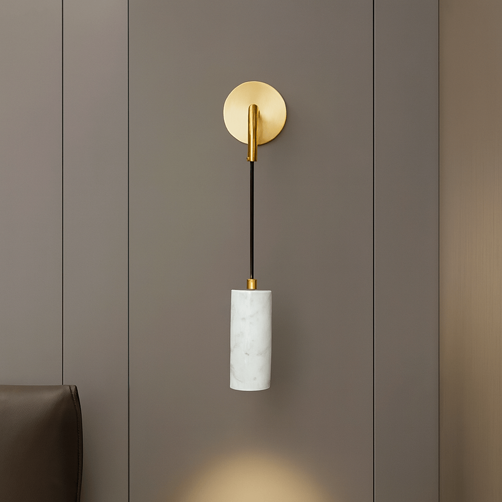 The Biltmore Marble Pendant Wall Sconce by Morsale, featuring a gold circular base, thin black stem, and natural marble shade, is mounted on a smooth gray wall with part of a brown chair visible in the lower left corner.