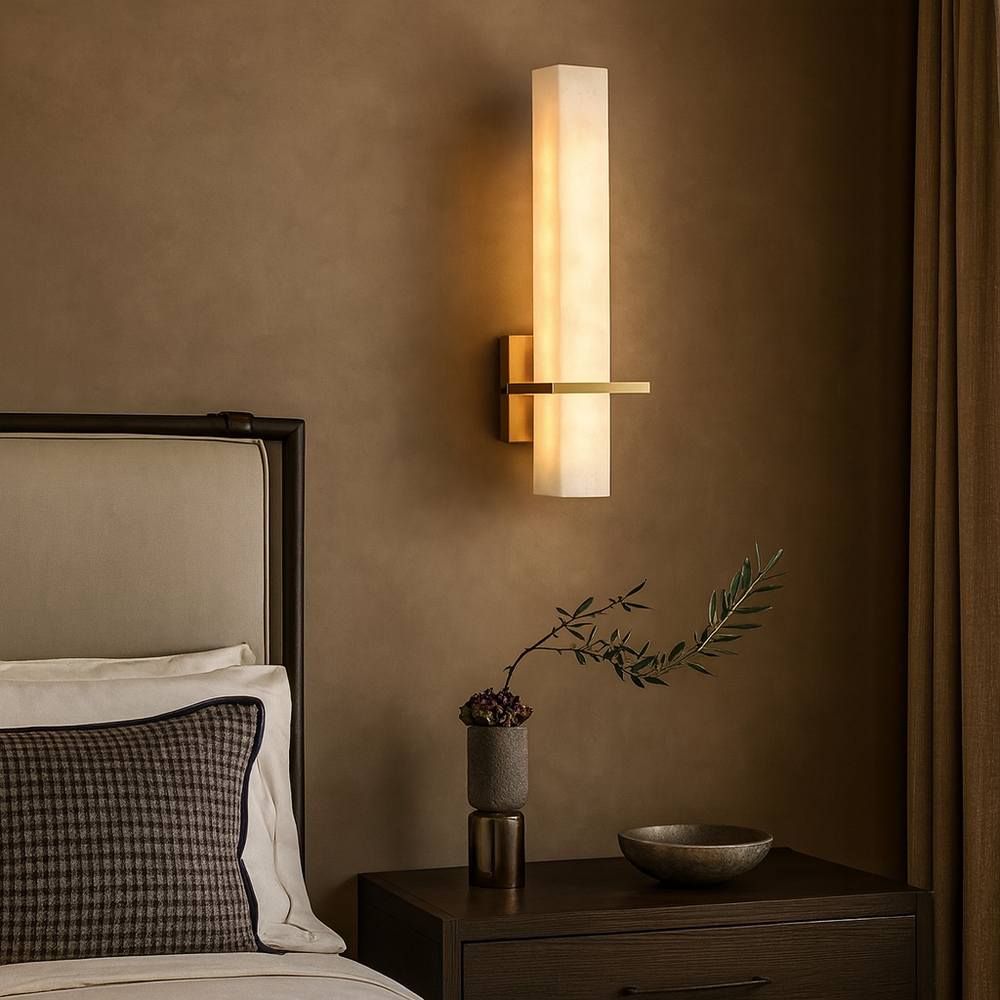 
                  
                    A Varella Natural Marble Wall Sconce by Morsale.com with a tall frosted light and brass accent is mounted above a dark nightstand holding a small vase, plant, and bowl next to a bed with checked pillow and upholstered headboard.
                  
                