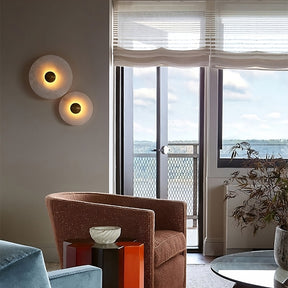 A cozy living room with a brown armchair, a blue sofa, and a small red side table with a decorative object. Two Moonshade Natural Marble Wall Sconces by Morsale.com featuring LED light sources add elegance to the space. A large window offers a scenic view of water and distant hills, with sheer curtains partially covering it.