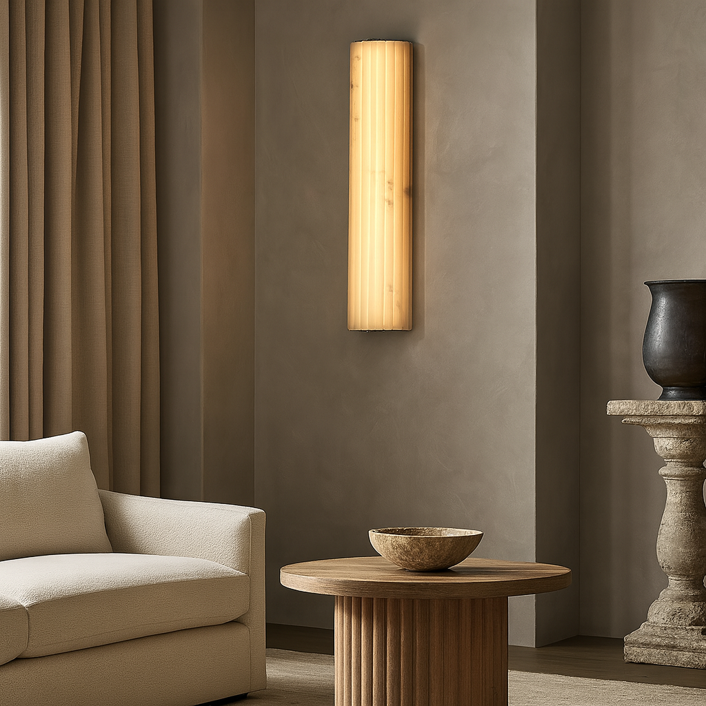 A cozy, modern living room with beige walls, a cream sofa, a round wooden coffee table, a tall sculpted vase on a pedestal, and the Morsale Marlo Marble Wall Sconce 18" glowing warmly on the wall.