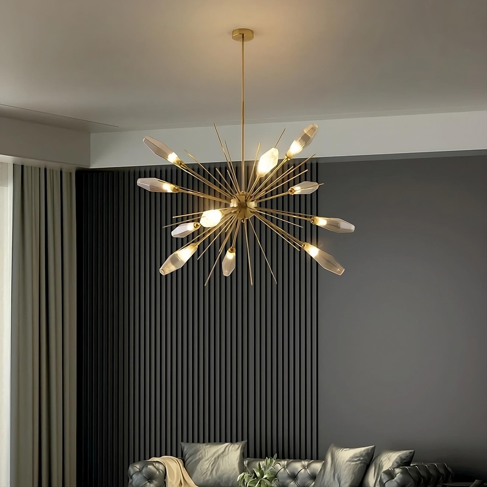 A modern living room features a stylish Morelli Sputnik Chandelier from Morsale.com with a starburst design, composed of brass rods and multiple exposed bulbs. The room has a dark accent wall with vertical textured panels, curtains, a tufted black sofa, and decorative pillows.