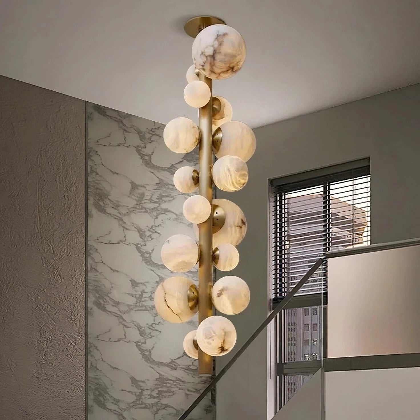 A Moonshade Luxury Marble Ball Chandelier by Morsale, featuring modern LED lights with a marble-like design, hangs in the stairwell. The walls display a textured pattern alongside a large window equipped with blinds. This elegant chandelier creates a striking contrast against the minimalist, gray interior.