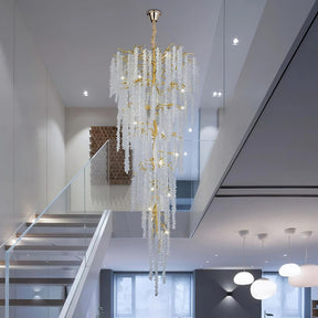 Modern interior featuring the Albero Crystal Foyer Chandelier by Morsale.com, a luxurious piece with cascading glass and polished gold accents, adorned with crystals hanging in a spacious, two-story room with minimalist design. A glass railing staircase is situated on the left and recessed lighting in the ceiling accentuates the chandelier’s elegance.