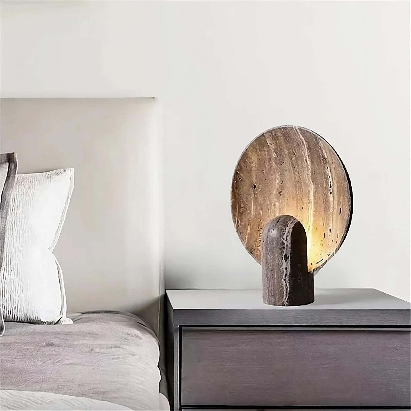 A modern bedroom features a gray cushioned headboard and white pillows on the bed. Beside the bed is a dark wooden nightstand with a unique circular Morsale Natural Travertine Table Lamp emitting warm light, creating a cozy atmosphere. The lamp's adjustable brightness adds to the room's vintage design charm.