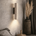 The Lunaro Travertine Wall Sconce by YEBMLP casts light up and down a textured beige wall, while a tall vase with pampas grass and dark drapes enhance the cozy, minimalist vibe of the softly neutral room.