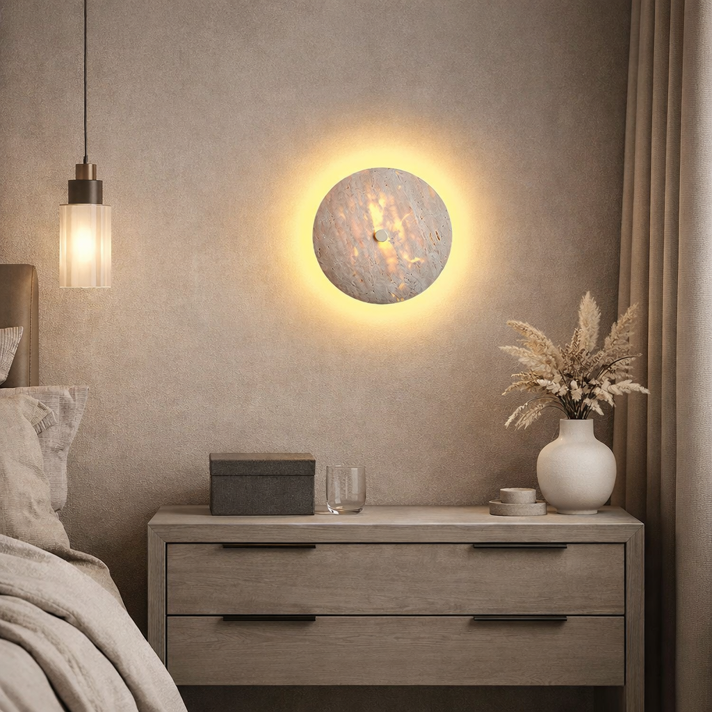 A cozy bedroom features the Morsale Brighton Travertine Wall Sconce glowing above a wooden nightstand with a vase of dried flowers, black box, and two glasses. A bed and modern hanging pendant light complete the minimalist look.