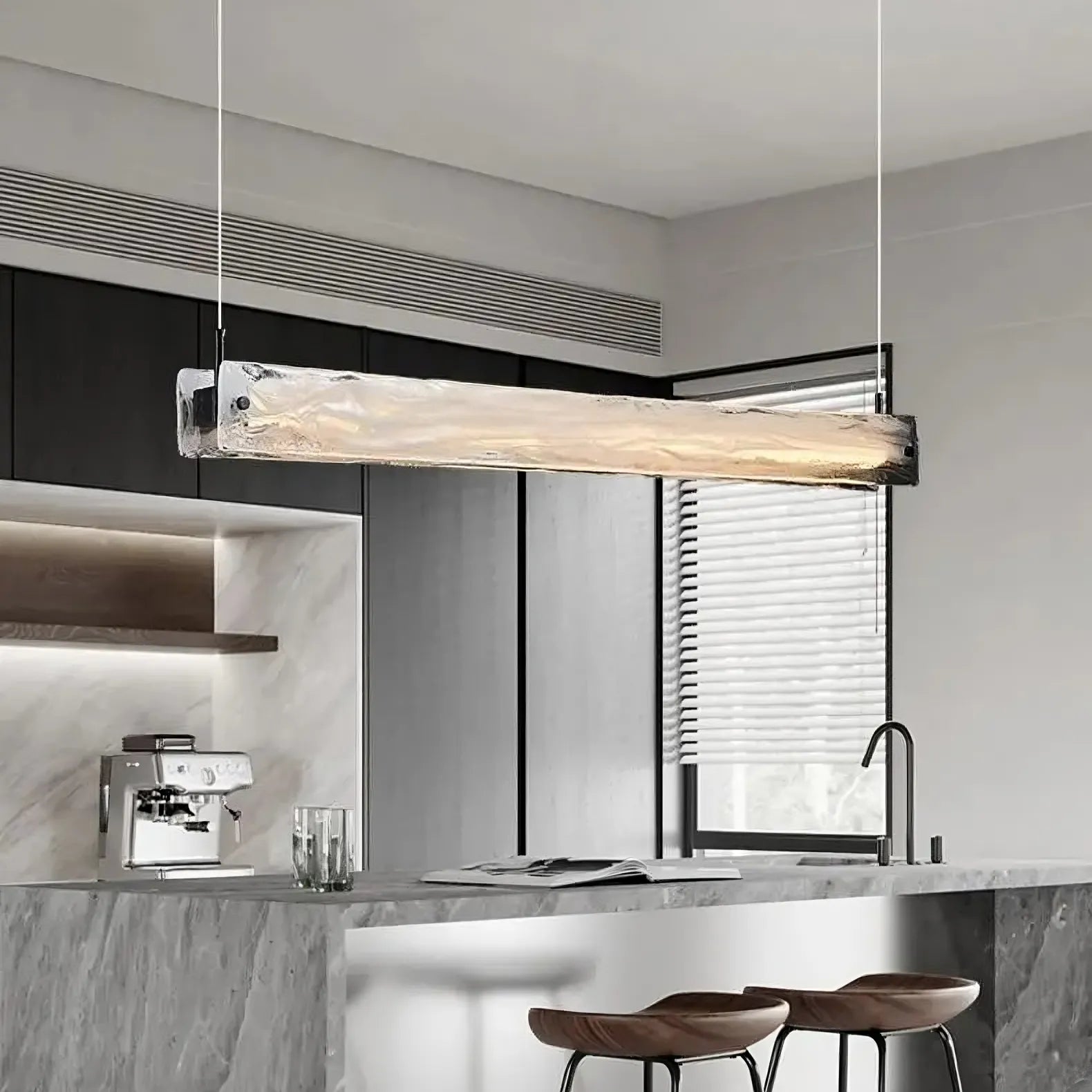 A modern kitchen with a marble countertop and backsplash showcases a sleek coffee machine. Two brown bar stools sit beneath the Costa Dining Room Linear Suspension Chandelier by Morsale, enhanced by subtle LED lighting. A window with blinds lets in natural light, completing the minimalist design.