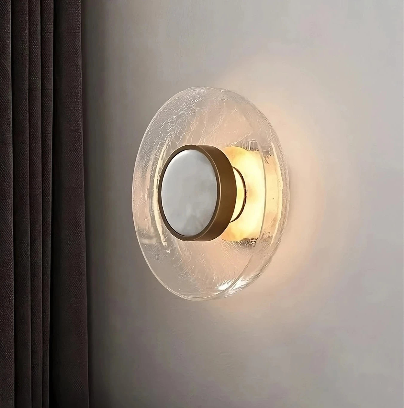 The Morsale Art Deco Cracked Glass Wall Sconce showcases a circular, crackled glass fixture with a central gold accent. It emits a soft, warm glow and is ideally mounted on a light-colored wall next to dark curtains.