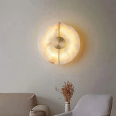 The Moonshade Natural Marble & Copper Wall Sconce by Morsale, featuring a circular design with a warm glow, is elegantly mounted on a beige surface above a gray armchair. A small bunch of dried flowers rests on a nearby table, enhancing the decor.
