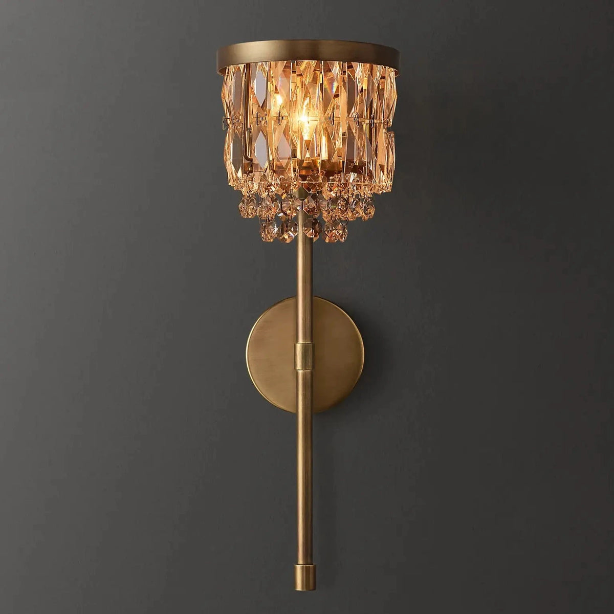 The Luxe Crystal Brass Wall Sconce from Morsale.com showcases a beautiful gold finish, complemented by a round backplate and a sleek vertical rod. Adorned with numerous crystal-like elements cascading from the top, this sophisticated lighting fixture enhances any room's decor, adding an opulent and refined atmosphere.