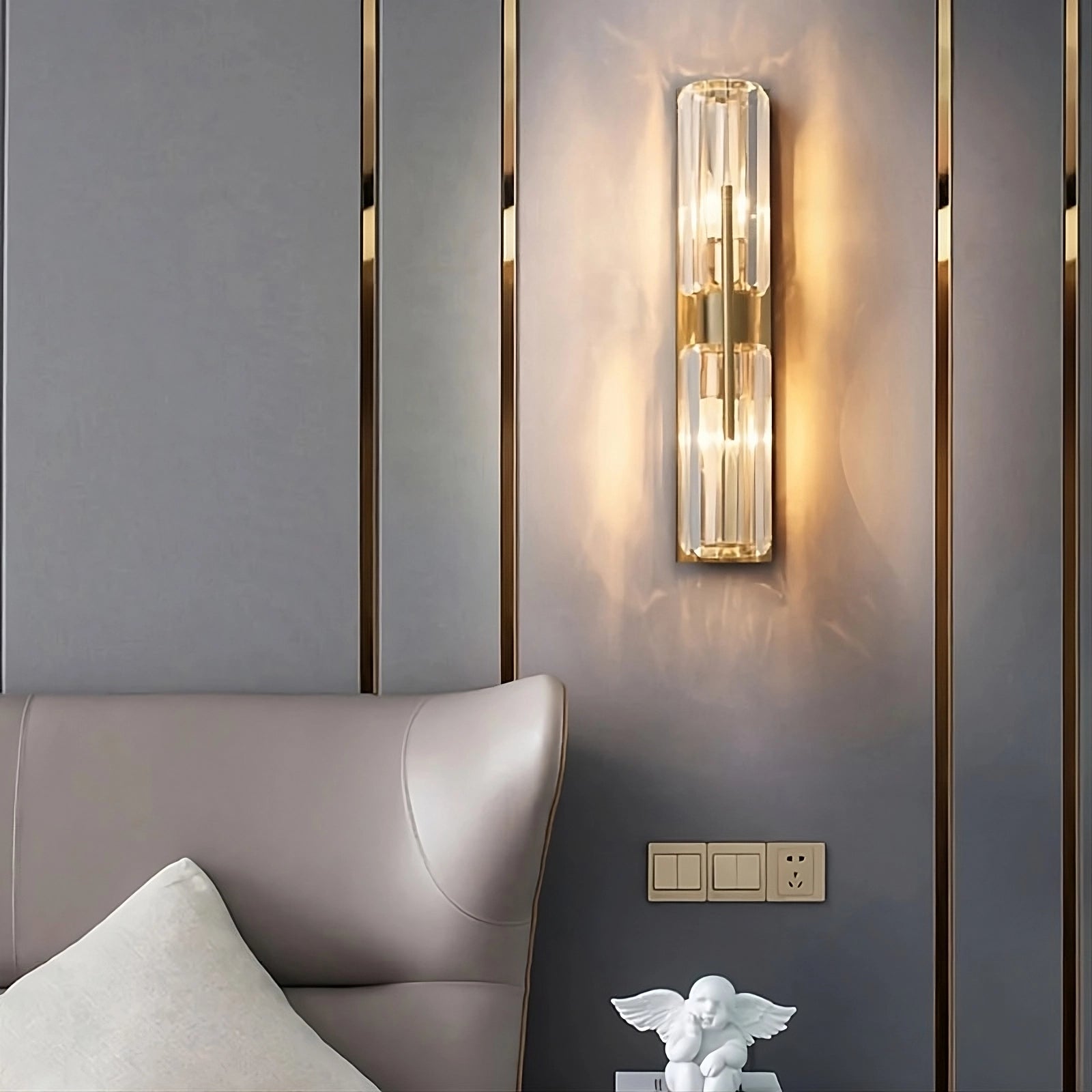 A bedroom showcases a modern style with the Morsale Gio Double Head Crystal Wall Sconce, emitting warm light and mounted on a gray paneled wall. Beneath it, two power outlets and a decorative white angel figurine add elegance. On the left side, you can see the edge of a cushioned headboard and pillow.