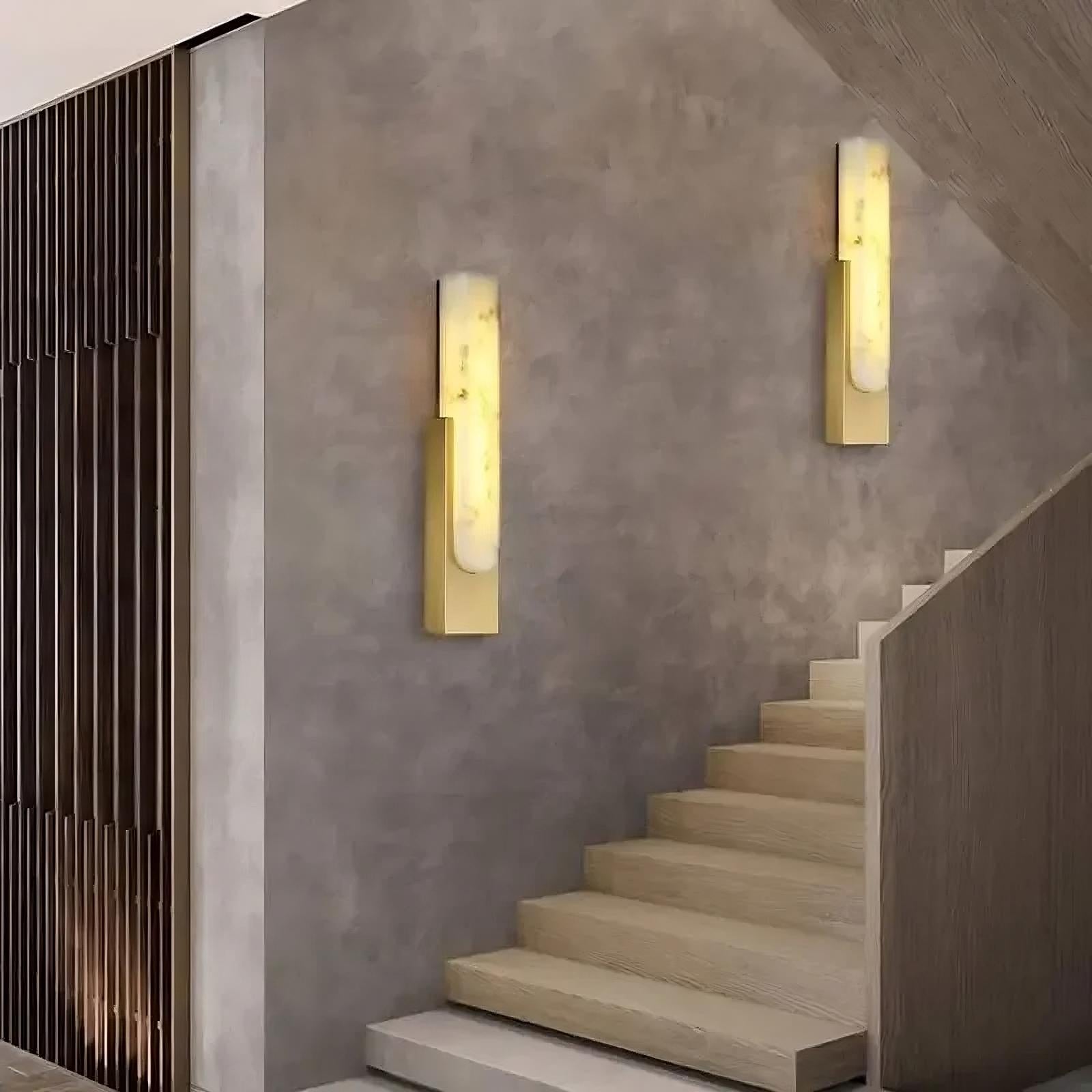 A modern, minimalistic staircase with light wooden steps and vertical wooden slats on the left wall. Two sleek, dimmable Moonshade Natural Marble Wall Sconces by Morsale.com, featuring a gold base and elongated frosted glass shades, are fixed on the gray concrete wall alongside the staircase.