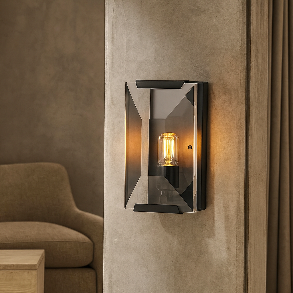 The Brookhill Crystal Wall Sconce from Morsale.com, featuring a glowing exposed bulb and angular iron frame, is mounted on a textured beige wall with an armchair and wooden table in the softly lit background.