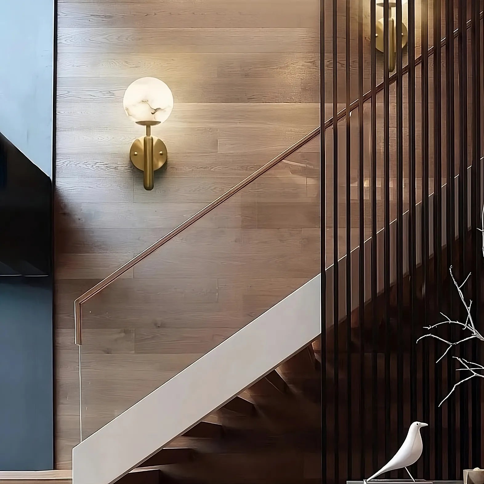 A modern staircase featuring a wooden accent wall, a sleek glass balustrade, and illuminated by stylish Natural Marble Sphere Wall Sconces from Morsale.com. A decorative white bird figurine sits at the base of the stairs next to a minimalist twig arrangement, adding unique texture to the design.