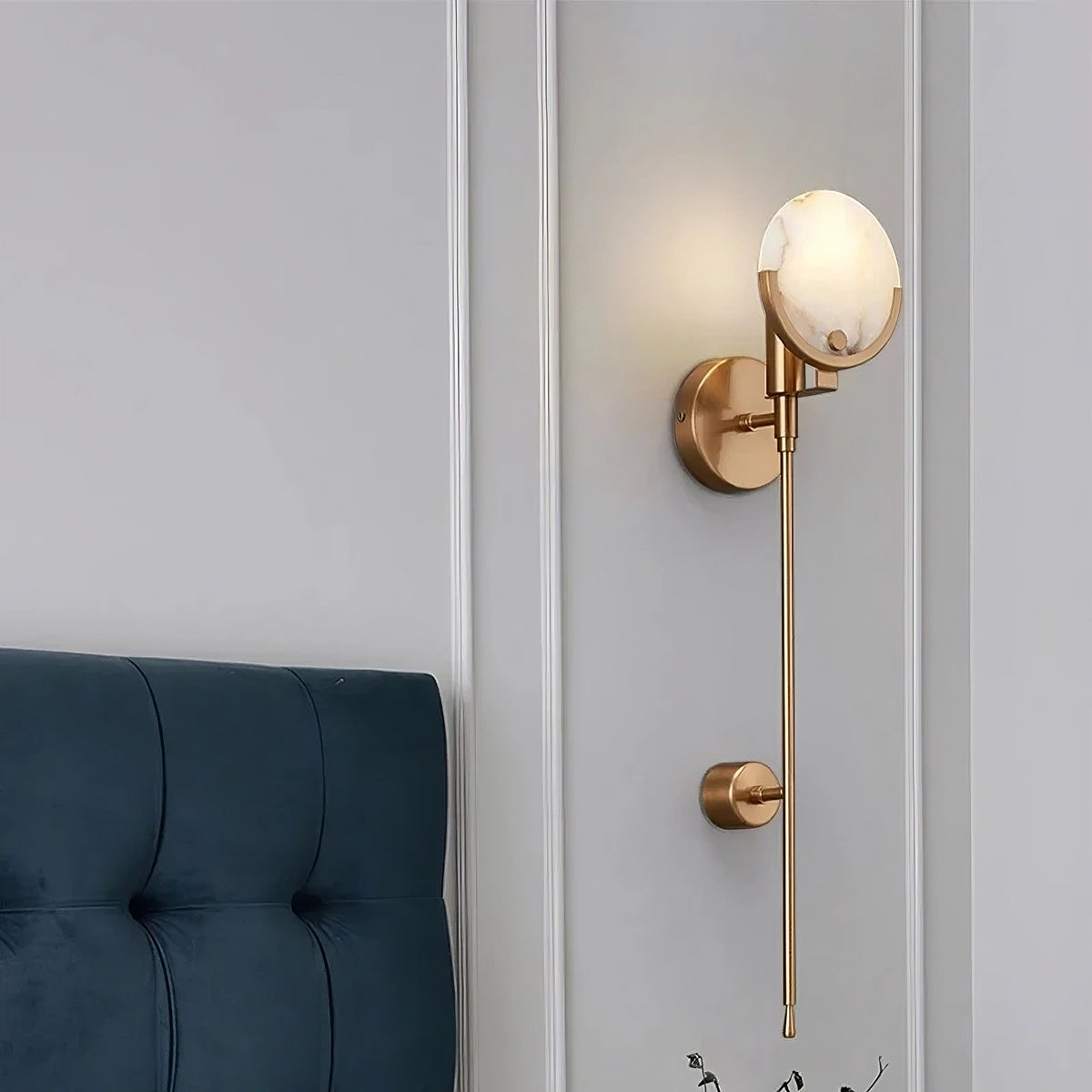 A Natural Marble Wall Light Sconce from Morsale.com with a brass finish and a round white lampshade is mounted on a light gray paneled wall. The lamp extends downward and is minimalist in design, embodying the elegance of unique marble patterns. A section of a dark blue tufted headboard is visible to the left.