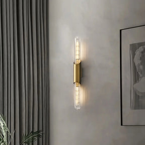 A Morsale Palermo 23" Brass Wall Sconce Light with two exposed bulbs is mounted on a light gray wall, embodying modern design. To the left, there are gray floor-to-ceiling curtains, and to the right, part of a framed black-and-white artwork is visible. A glimpse of green foliage at the bottom left adds an elegant touch.