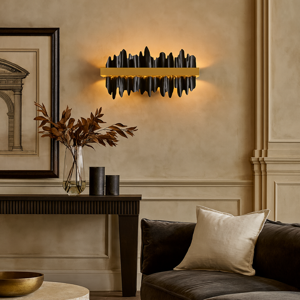 A modern living room features the sculptural black Nera Wall Sconce by Morsale.com emitting warm light, a sleek black console table with a vase of dried leaves, two black cylinders, framed art, and a dark sofa with a cream pillow.