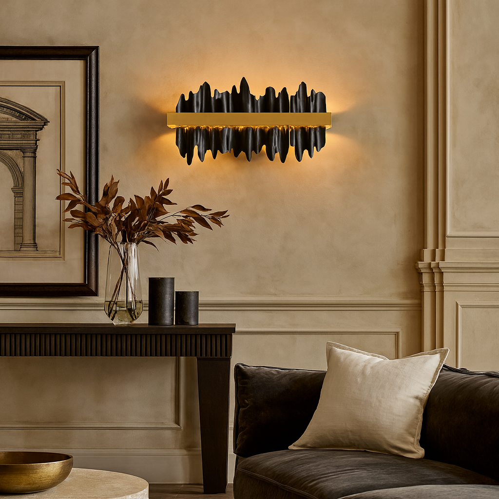 A modern living room features the sculptural black Nera Wall Sconce by Morsale.com emitting warm light, a sleek black console table with a vase of dried leaves, two black cylinders, framed art, and a dark sofa with a cream pillow.