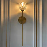 The Clear Crystal & Brass Wall Light Fixture by Morsale.com features a cylindrical brass stem with a round base mounting. This light, adorned with clear crystal and brass elements, is topped with a faceted glass orb that emits ambient lighting. It casts a warm glow against a minimalistic, pale grey wall adorned with vertical gold lines.