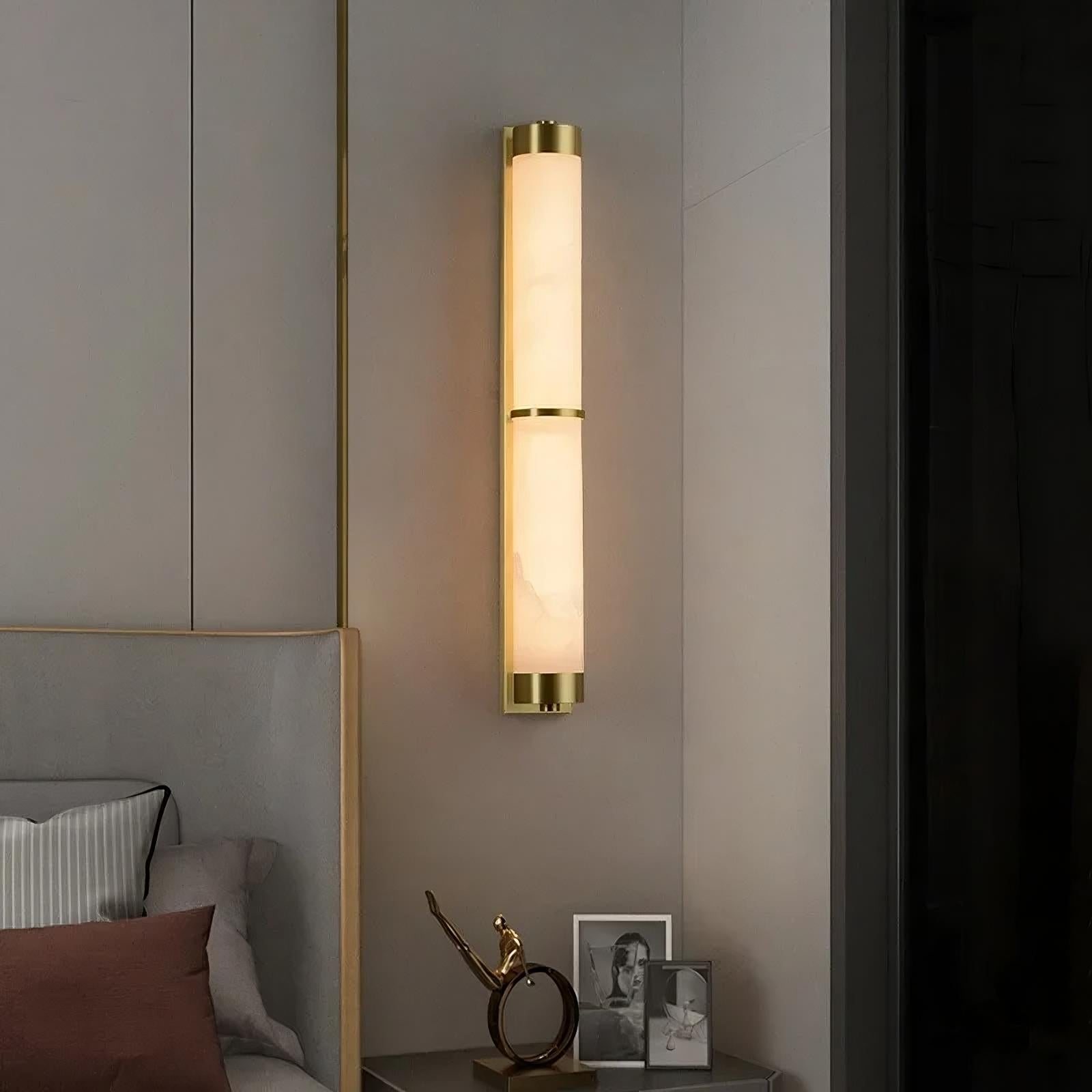 A modern bedroom features a sleek, cylindrical wall-mounted light fixture with a brass finish and frosted glass. The 23" Natural Marble & Brass Wall Light from Morsale illuminates the space elegantly. On a small bedside table below, there are two framed photos and a decorative sculpture of a gold-toned figure in a dynamic pose.