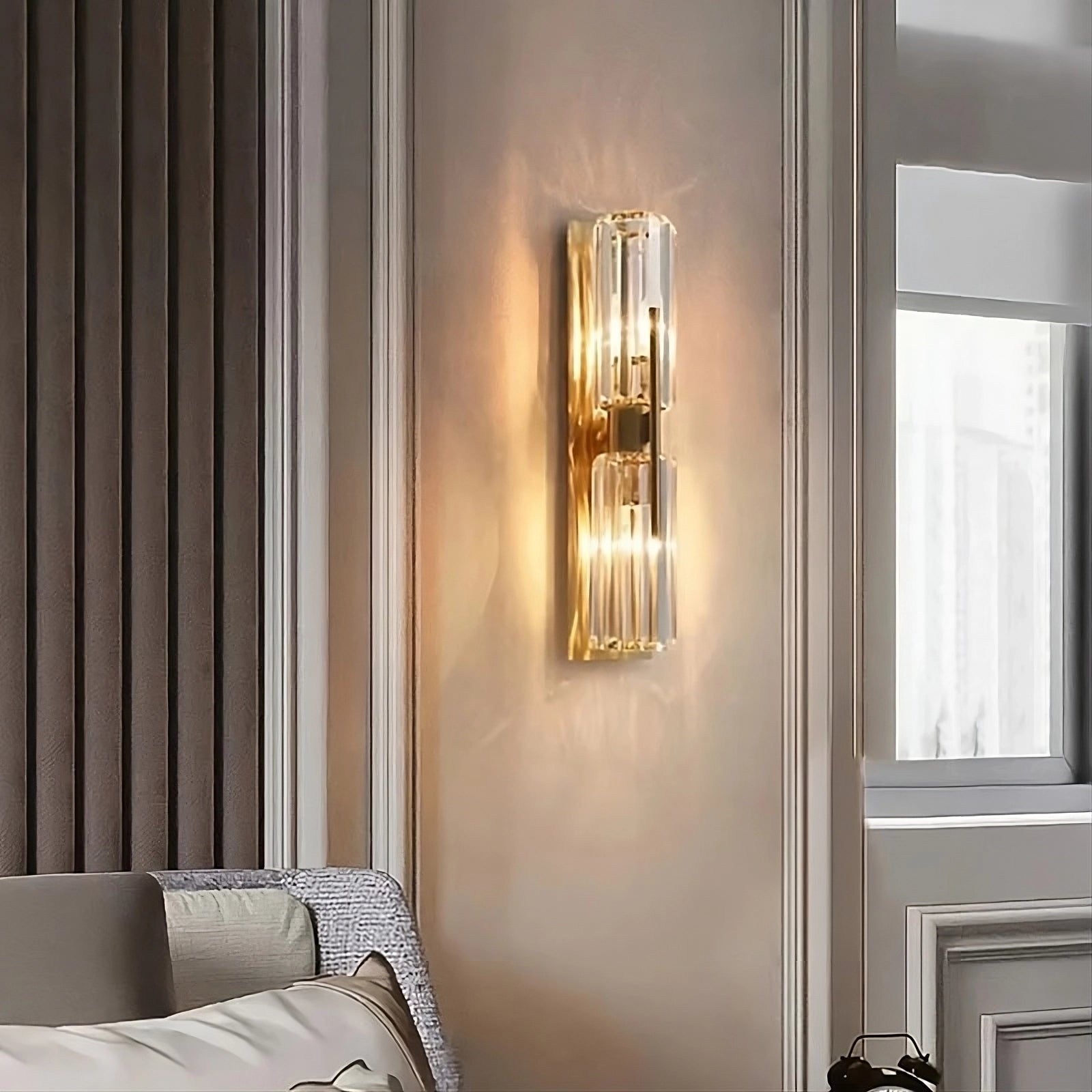 The Gio Double Head Crystal Wall Sconce by Morsale features a gold metal frame and clear glass covering, casting warm LED lighting against a beige wall. Below, there are white and green flowers.
