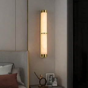 A modern bedroom features a sleek, cylindrical wall-mounted light fixture with a brass finish and frosted glass. The 23" Natural Marble & Brass Wall Light from Morsale illuminates the space elegantly. On a small bedside table below, there are two framed photos and a decorative sculpture of a gold-toned figure in a dynamic pose.