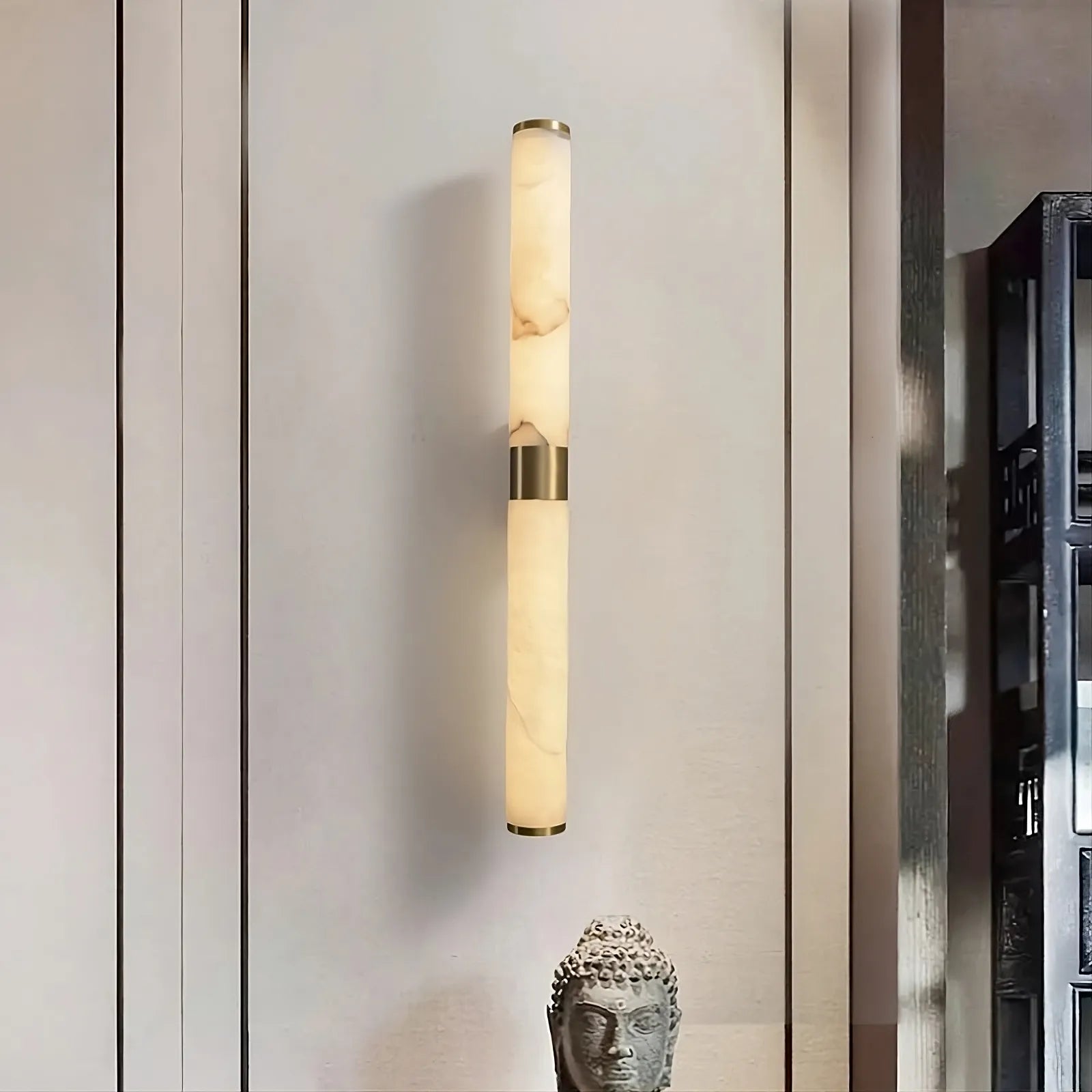 A 27" Moonshade Natural Marble & Copper Wall Sconce by Morsale emits a soft, warm glow as it's mounted on a light-colored wall. Below it, a small decorative sculpture of a serene face enhances the contemporary home décor with an artistic flair.