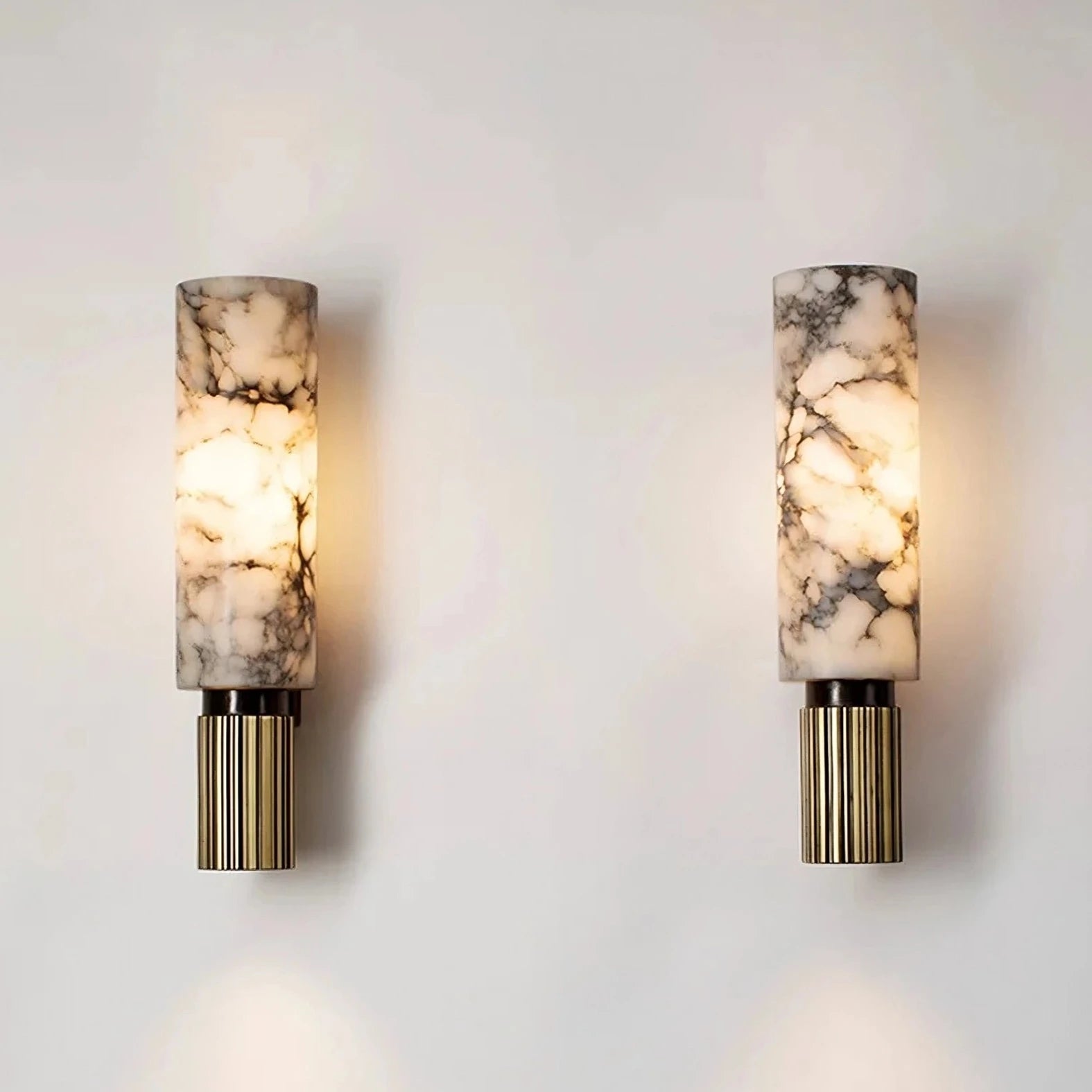 A pair of stylish, handmade Moonshade Natural Marble Sconces from Morsale.com featuring cylindrical, marble-patterned lampshades and ribbed golden bases emit a soft, warm light from dimmable LED bulbs, mounted on a plain white wall. The elegant design provides a modern and sophisticated look perfect for natural marble decor.