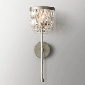 The Luxe Crystal Brass Wall Sconce by Morsale.com showcases a round backplate and an elongated arm in a striking metallic finish. This exquisite fixture boasts a cylindrical shade adorned with dangling crystals, casting a soft and luxurious glow against the plain gray wall. It's the perfect addition to any luxury home decor.