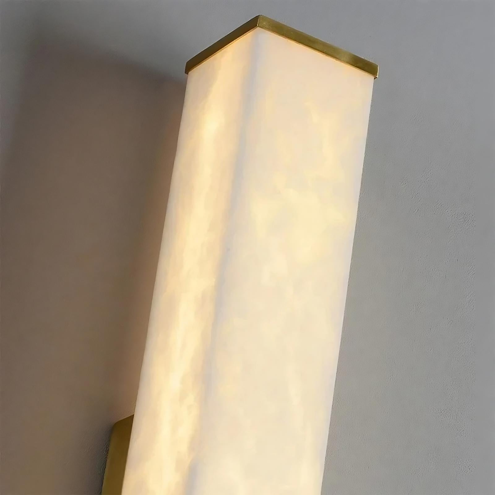 A close-up view of a wall-mounted rectangular Natural Marble Indoor Wall Sconce Light with a frosted white cover and brass-colored trim against a light grey wall. The light illuminates with a soft, diffused glow, highlighting the minimalistic and modern design from Morsale.com.