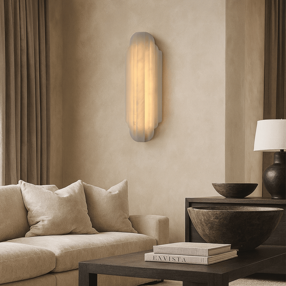 
                  
                    A modern living room with a beige sofa, neutral pillows, a dark coffee table with stacked books and bowls, beige walls, an Arina Marble Sconce by Morsale casting a warm glow, and floor-length curtains.
                  
                