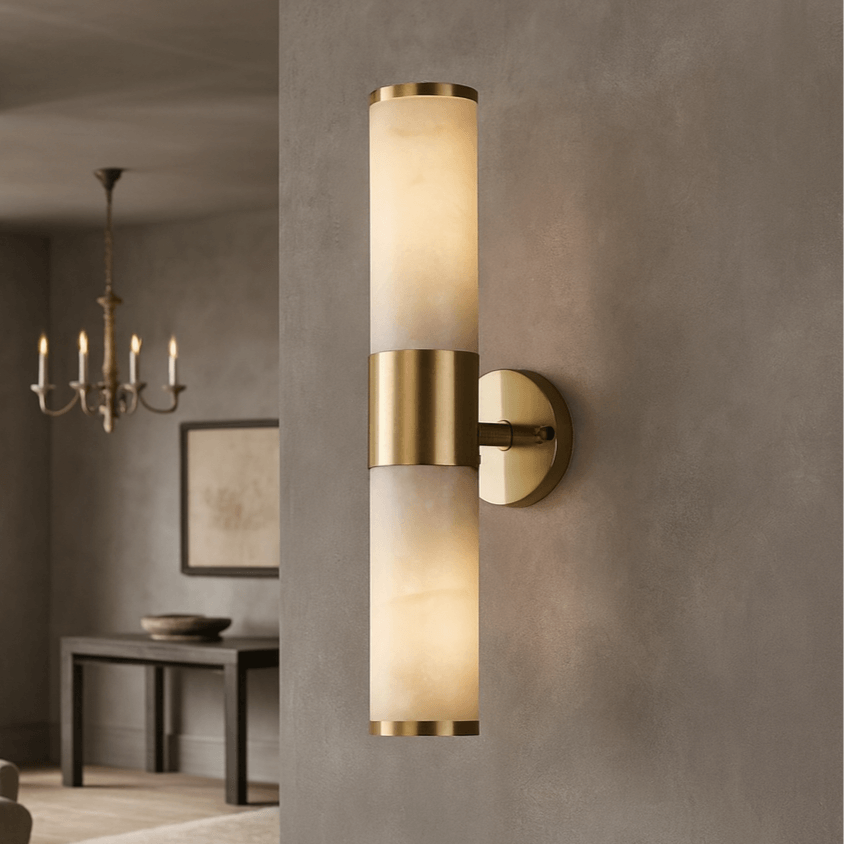 The Aurelia Natural Marble Wall Sconce by Morsale.com features two cylindrical frosted glass shades, brushed brass accents, and solid brass hardware, mounted on a gray wall with a dining area and art visible in the background.