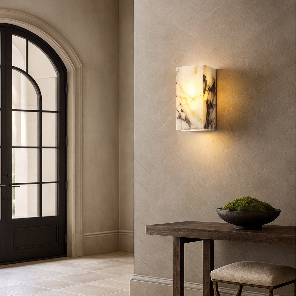 The Morsale Arden Marble Wall Light casts a warm glow in an elegant entryway with beige textured walls, a dark wood console, a moss-filled decorative bowl, tiled flooring, and a large black-framed glass door.
