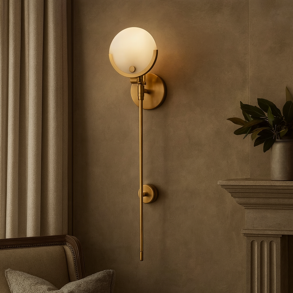 The Terra Marble Wall Sconce by Morsale.com features a gold natural marble base, round frosted glass shade, and integrated LED, casting warm white light in an elegant living room beside gray curtains, a plant, and a fireplace mantel.